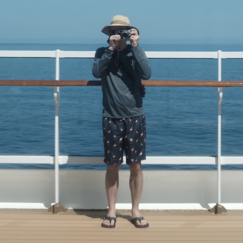 A man, wearing a blue shirt and a brown hat, holding a camera, standing on the deck of a ship, the ocean behind him, taking a photograph of his own reflection in a window.
