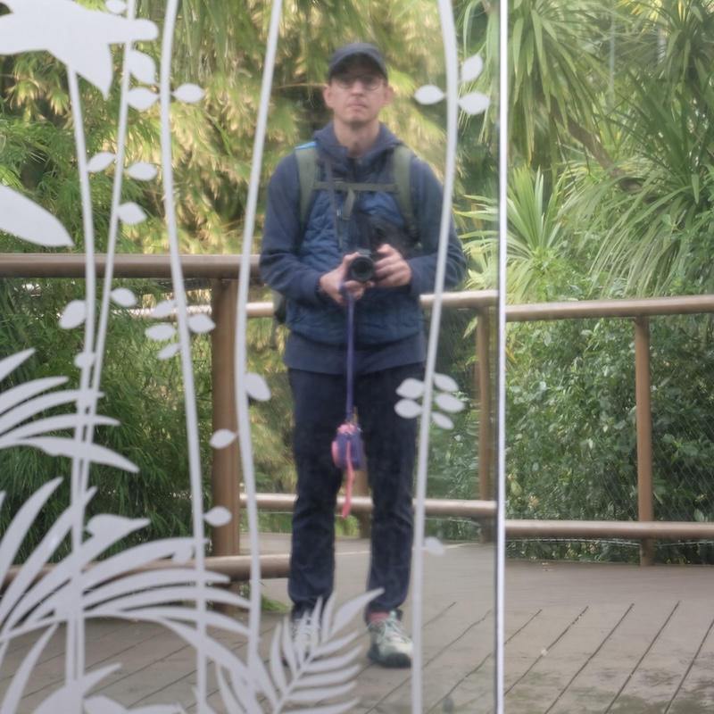 A man, wearing a blue vest and a blue hat, holding a camera, standing in front of small trees and plants, taking a photograph of his own reflection in a glass door.