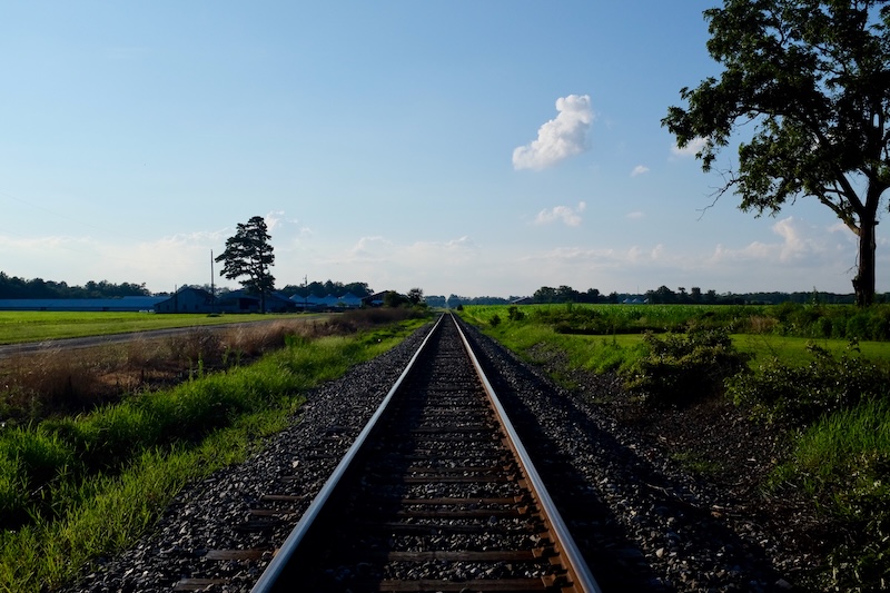 Train tracks looking north, disappearing in the distance with gray stones and green grass on either side, trees and farm buildings near the horizon.
