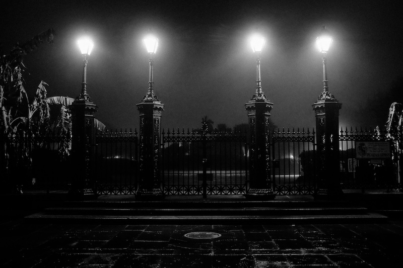 Four large lamps above the closed gate, along the fence outside of Jackson Square, their light dispersing into the evening fog.