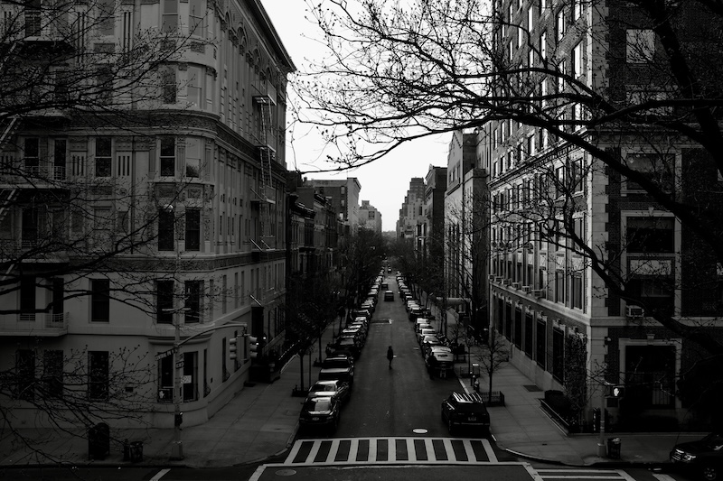 A black and white photograph of a city street, cars and buildings lining both sides. A person crosses the street and appears to pause in the middle.