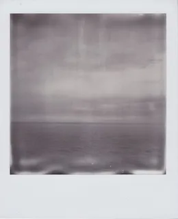 A monochrome Polaroid photograph of a cloudy sky over the Atlantic Ocean, a buoy floating in the distance.