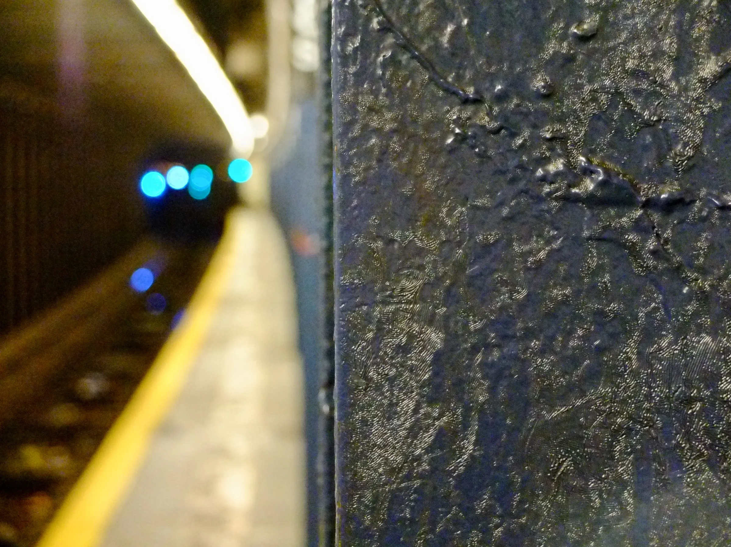 Blue paint caked onto vertical steel beams that support a subway platform, down the tunnel, lights from an out of focus train that approaches