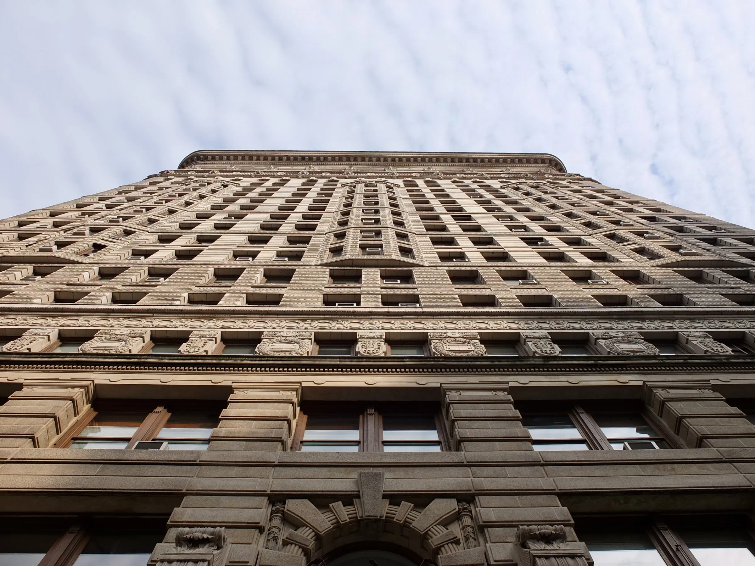 Pillowy clouds cover a blue sky above the west facade of the Flatiron building. Looking up the structure is filled with ornate details: carved stone, patterned brickwork, protruding bay windows.