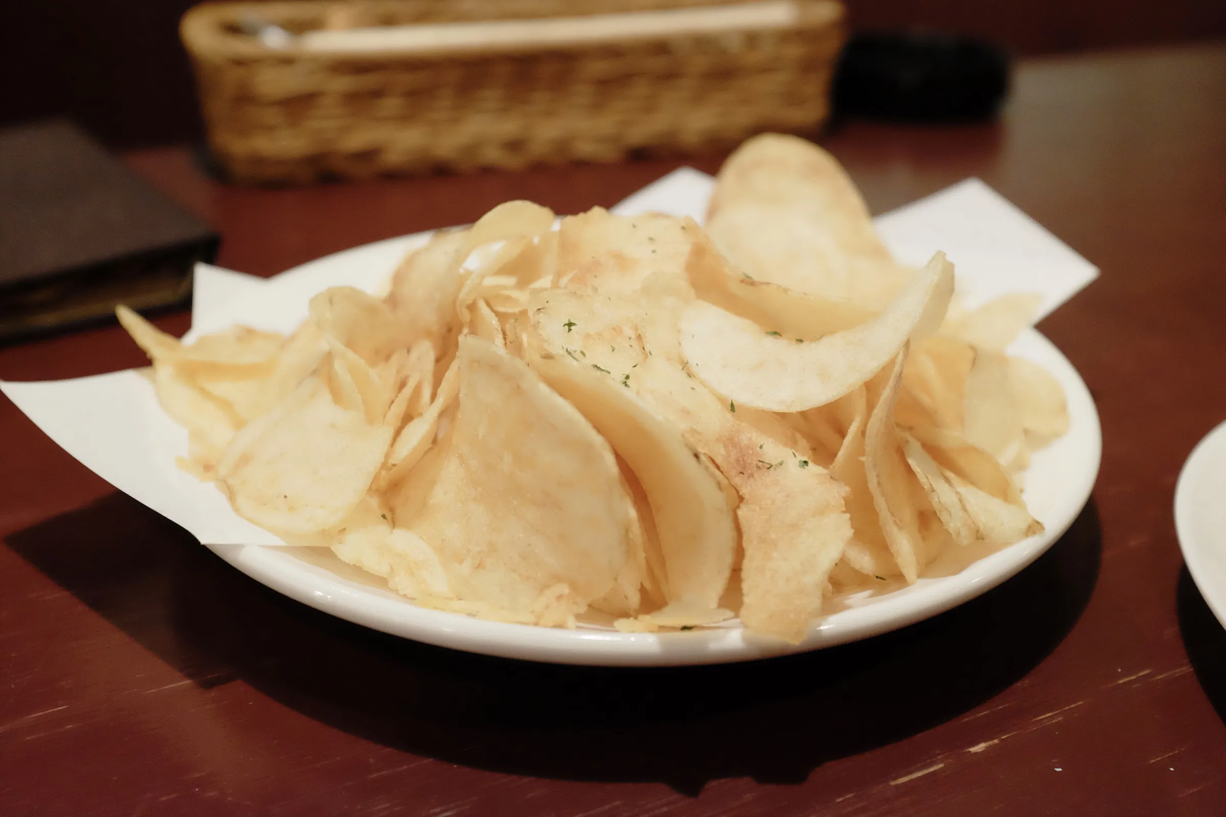 Golden yellow potato chips, dusted with bright green flecks of dill, sit on a white plate that rests on a wooden bar.