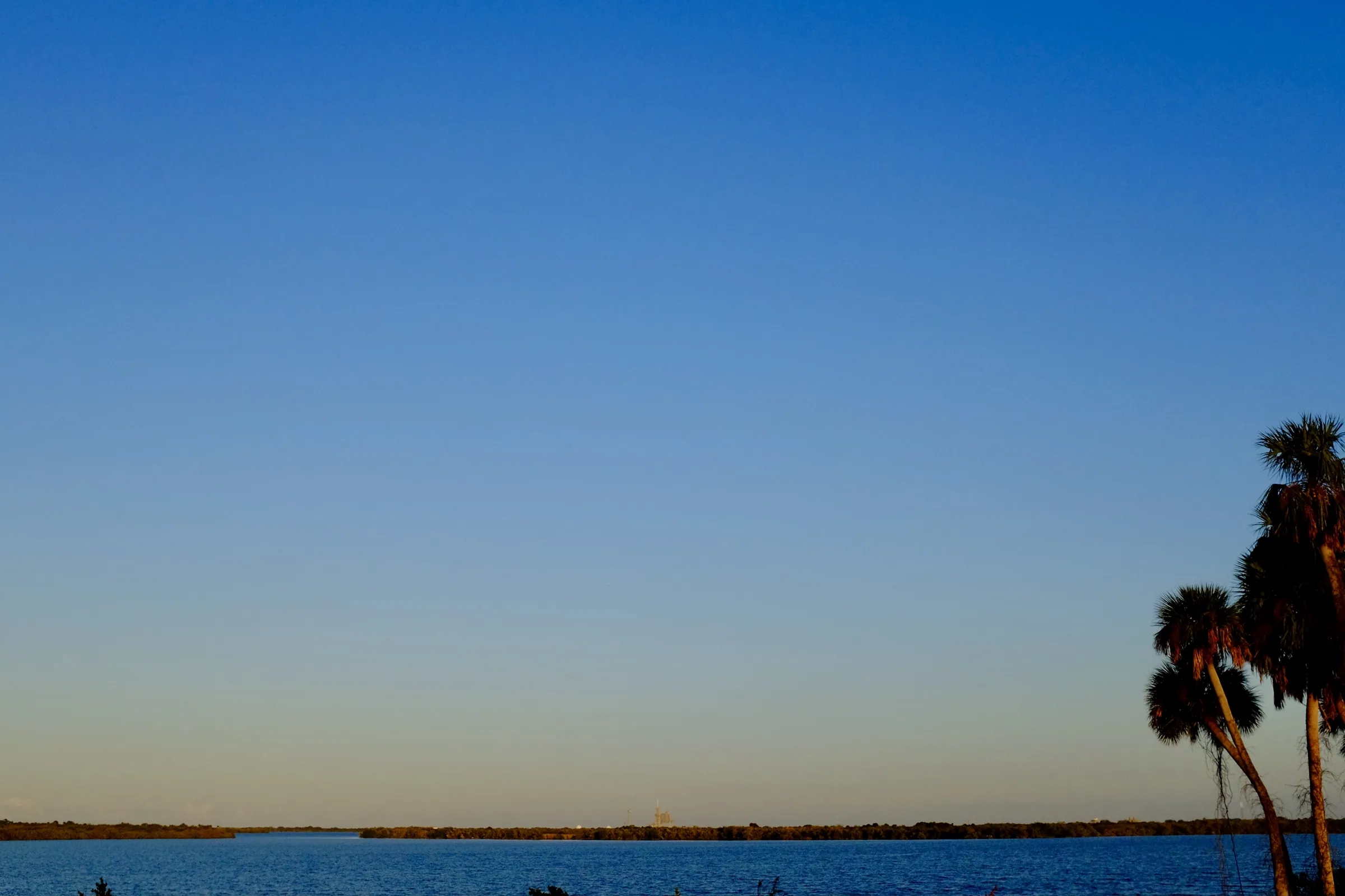 Palm trees and water, looking toward Cape Canaveral in the distance where a rocket, not destined to launch this day, sits waiting in front of a blue sky, beginning to turn pale orange in the fading light.