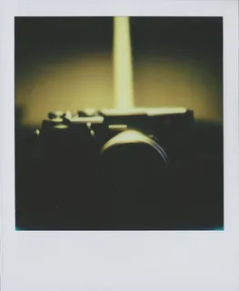 A color Polaroid photograph of a silver and black camera, a Fuji X100S, the top of camera reflecting pale yellow light, out of focus.