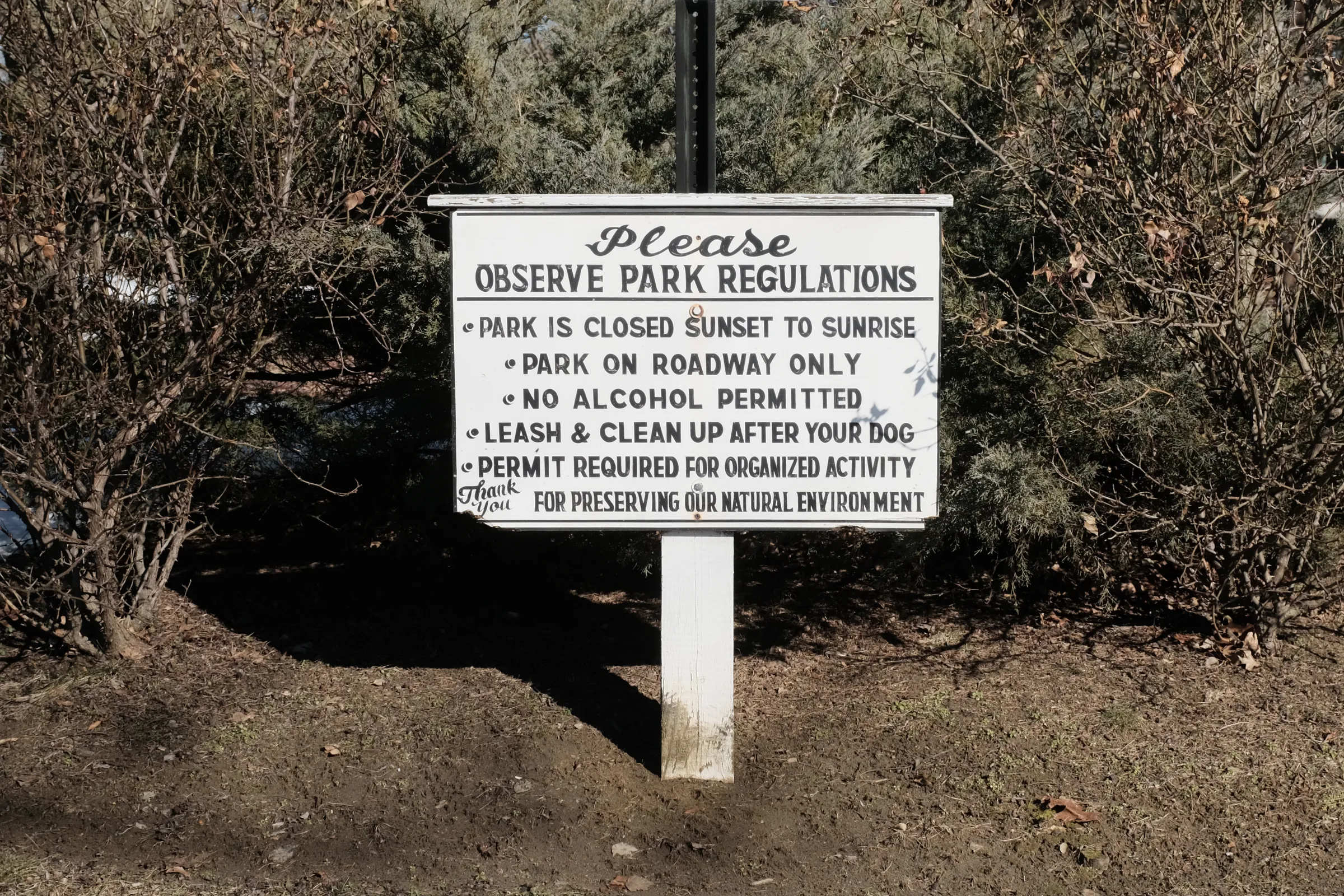 A small rectangular, wooden sign on a wooden post. The white paint is beginning to chip, the black letters are faded. The words appear hand stenciled, except for wide script of 'please' and 'thank you.' The sign reads: 'Please, observe park regulations. Park is closed sunset to sunrise. Park on roadway only. No alcohol permitted. Leash and clean up after your dog. Permit required for organized activity. Thank you for preserving our natural environment.'