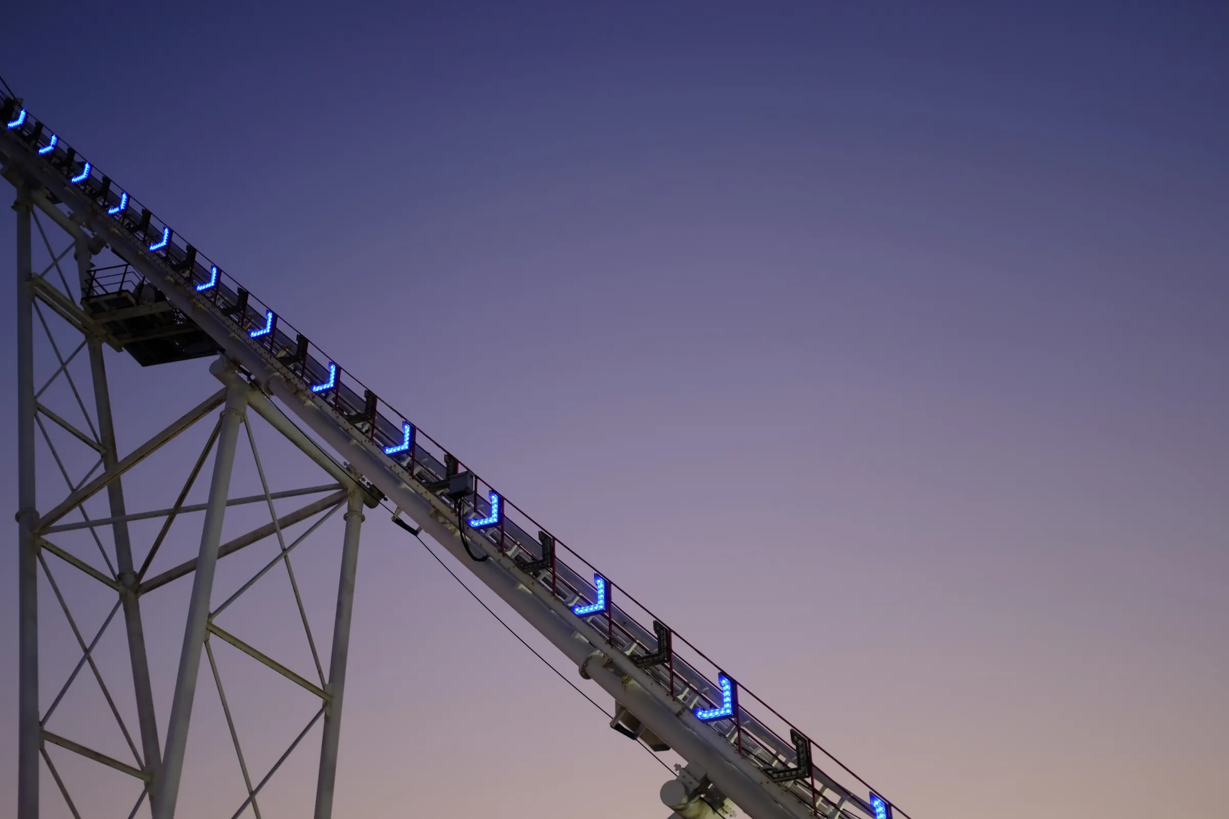 A roller coaster track, descending downward at 45 degrees, blue arrows lets along the line, in front of purple evening sky blending into the last bit of pastel orange from the setting sun.