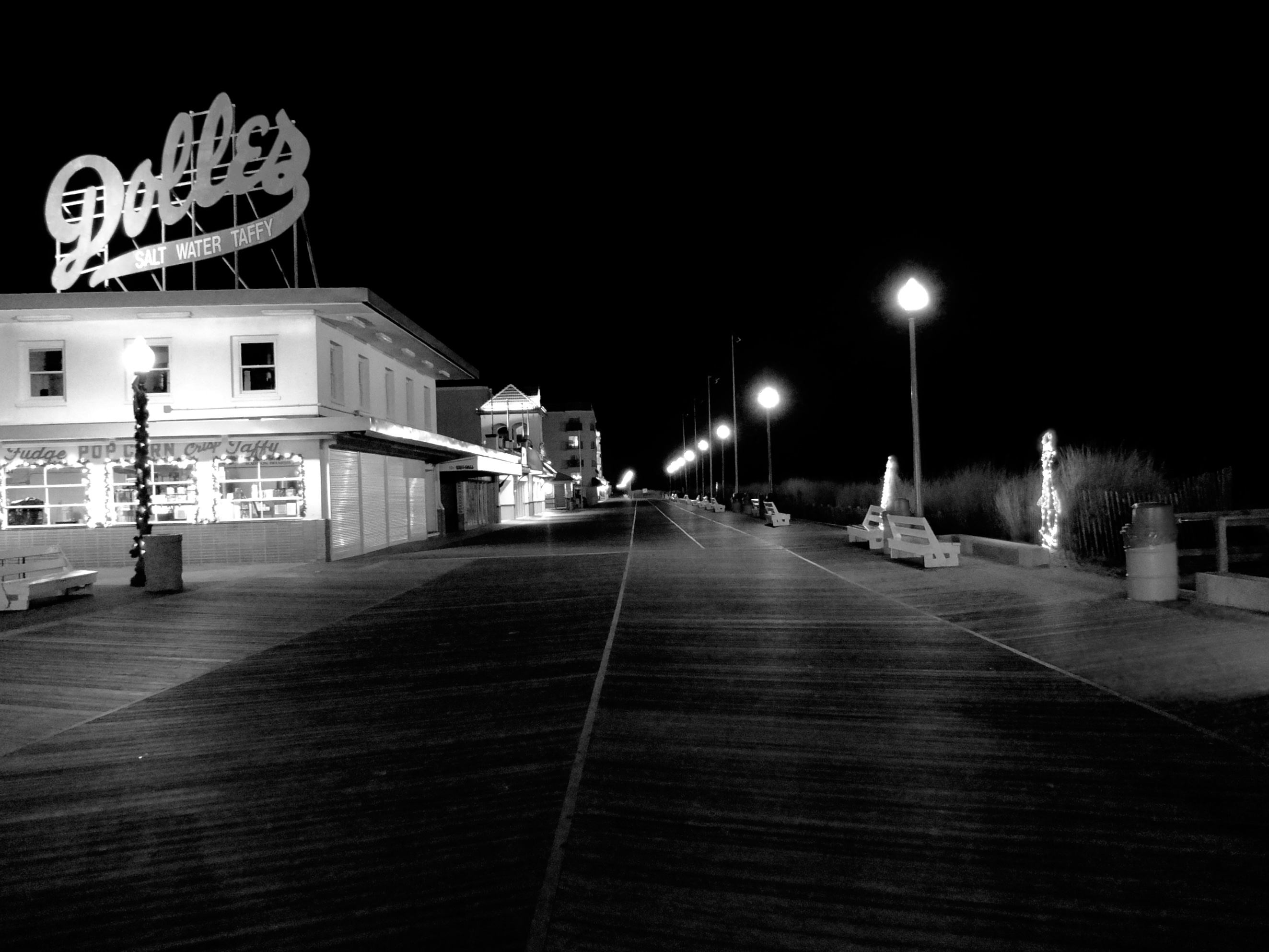 An empty boardwalk at night, street lamps along the beach edge, a large sign above a two-story building sign reads Dolles.
