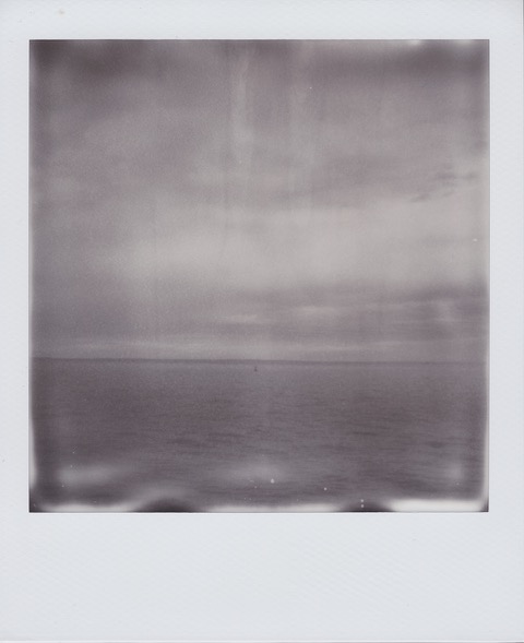 A monochrome Polaroid photograph of a cloudy sky over the Atlantic Ocean, a buoy floating in the distance. Photo taken in Brooklyn, New York City, USA.