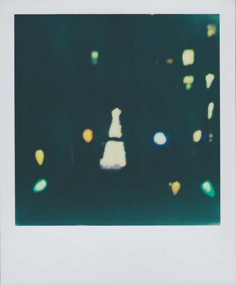 A color Polaroid photograph of a white, brightly lit Christmas tree, colored lights surrounding it, out of focus. Photo taken in Manhattan, New York City, USA.