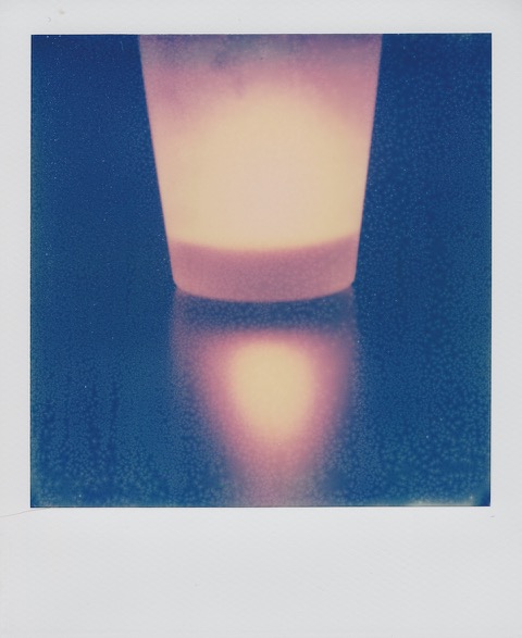 A color Polaroid photograph of a small white candle and its yellow flame, reflecting off the surface of a dark table. Photo taken in Queens, New York City, USA.