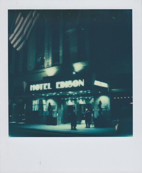 A monochrome Polaroid photograph of hotel entrance. Doormen stand on the sidewalk outside. A large American flag hangs from a flagpole above. A large sign on the hotel reads 'Hotel Edison' in Art Deco letterforms. Photo taken in Manhattan, New York City, USA.