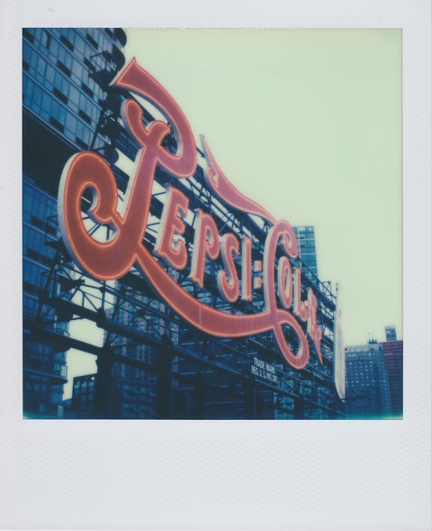 A color Polaroid photograph of a large, red Pepsi-Cola sign held up by a metal scaffolding structure. In the background, several large buildings. Photo taken in Queens, New York City, USA.