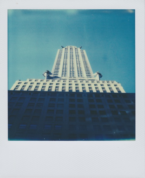A color Polaroid photograph looking up at a tall, gray, Art Deco building. The bottom of the building is darkened by a shadow, the top a brighter gray. Bright blue sky in the background. Photo taken in Manhattan, New York City, USA.