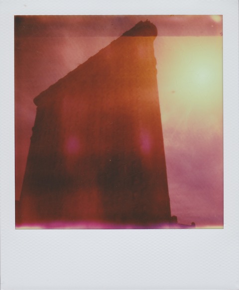 A color Polaroid photograph of the silhouette of a large building. On the right, a bright yellow sun shines in the sky. Photo taken in Manhattan, New York City, USA.