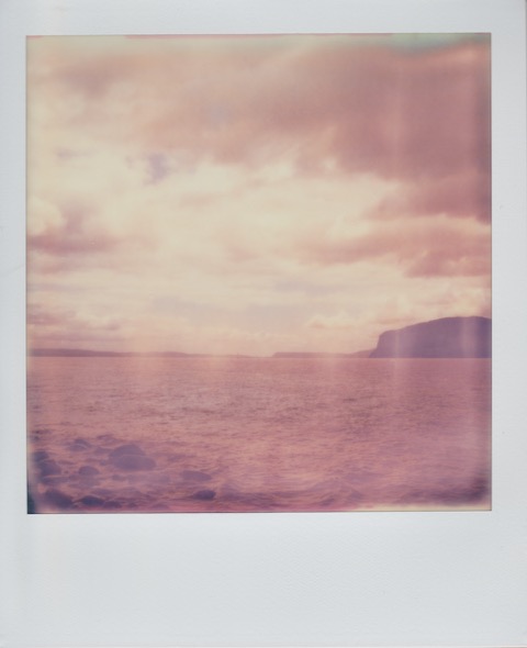 A color Polaroid photograph of water. Above, gray and white clouds and the sky. In the distance, on the right, a tall, rocky piece of land. Photo taken in Newport, Rhode Island, USA.