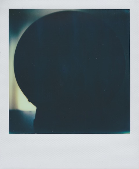 A color Polaroid photograph of a clock. Frame is dimly lit, on the silhouette, and really only the left side. Photo taken in Queens, New York City, USA.