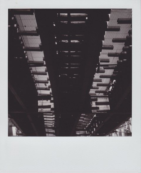 A monochrome Polaroid photograph of the underside of raised train track, bright sunlight blocked by the repeating pattern of railroad ties. Photo taken in Queens, New York City, USA.