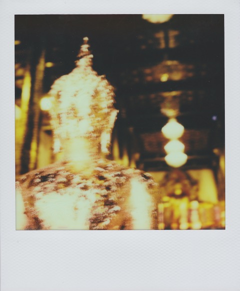 An out-of-focus color Polaroid photograph of gold Buddha statue (seated, in the Thursday, Pang Samti, position). In the background, the interior of a temple, bright overhead lights. Photo taken in Bangkok, Thailand.