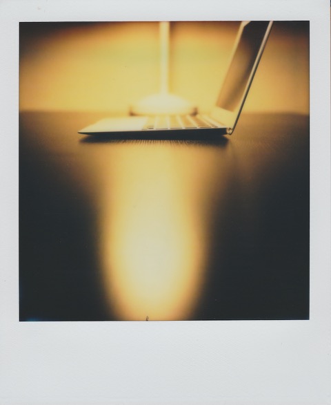A color Polaroid photograph of an 11-inch Apple Macbook Air, sitting on a desk. Behind it a lamp, reflecting off the table. Photo taken in Queens, New York City, USA.