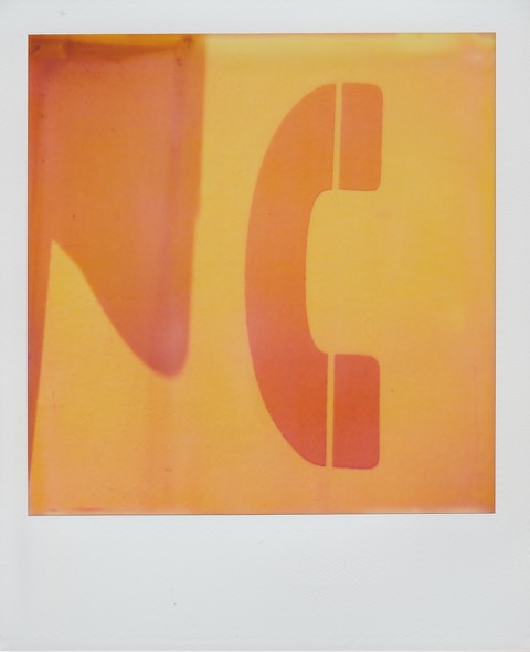 A color Polaroid photograph of the outside of a bright yellow telephone box, a large orange, or darker yellow, phone symbol on the side. Photo taken in Queens, New York City, USA.