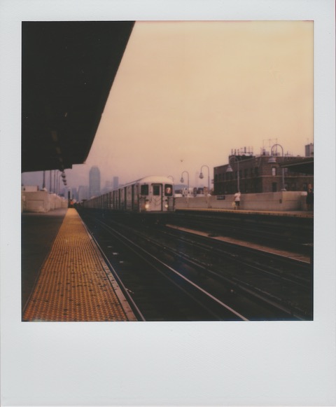 A color Polaroid photograph of eleveated rail track. A 7 train on the right side of tracks. In the distance, Manhattan. On the left, along the tracks, yellow flooring. Photo taken in Queens, New York City, USA.