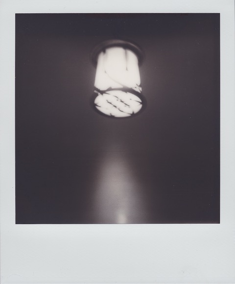 A monochrome Polaroid photograph of cylindrical ceiling lamp hanging above a building entryway. Photo taken in Queens, New York City, USA.