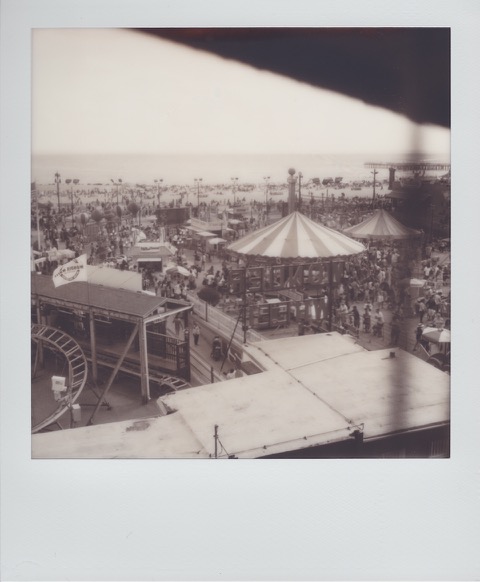 A monochrome Polaroid photograph of a crowded boardwalk and beach. Taken from some height above, in the foreground, the tops of large, striped tents. In the distance, the ocean. Photo taken in Brooklyn, New York City, USA.