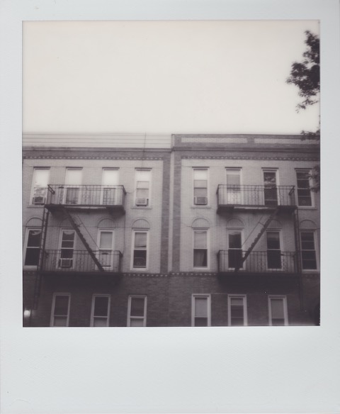A monochrome Polaroid photograph of two, three-story brick and masonry apartment buildings, joined at the edge. A separate, iron fire-escape hangs from each building. Photo taken in Queens, New York City, USA.