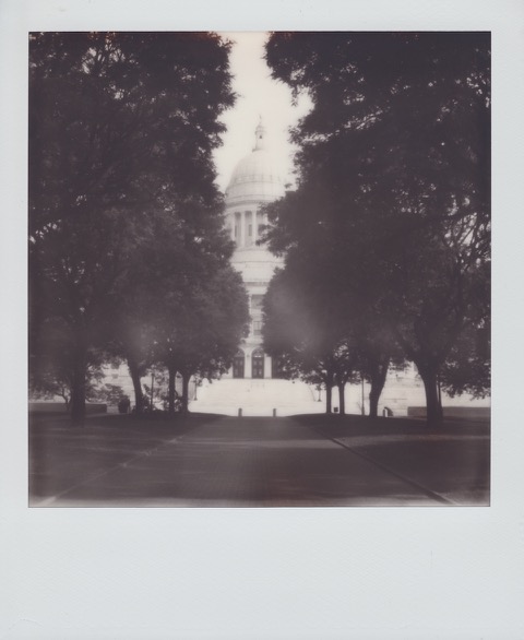 A monochrome Polaroid photograph of a park. On the sides, large trees. In the distance, a large, neoclassical state capitol building. Photo taken in Providence, Rhode Island,  USA.