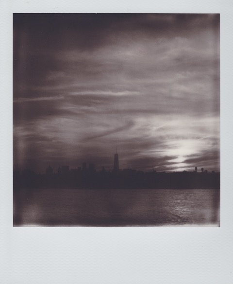 A monochrome Polaroid photograph of a river. In the distance, a city skyline with many tall buildings and one very tall building. Photo taken in Anytown, New Jersey, USA.