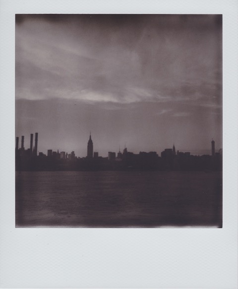 A monochrome Polaroid photograph of a river with a city skyline silhouette in the distance. The Empire State Building clearly visible. Photo taken in Queens, New York City, USA.
