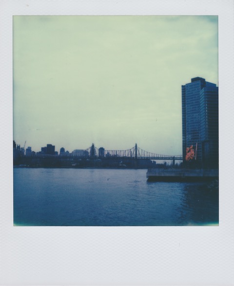 A color Polaroid photograph a river. A large neon side on the right bank and a bridge in the distance. Photo taken in Queens, New York City, USA.