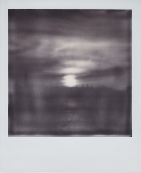 A blurry monochrome Polaroid photograph of a sun setting, a river and city skyline in the distance. Photo taken in Anytown, New Jersey, USA.
